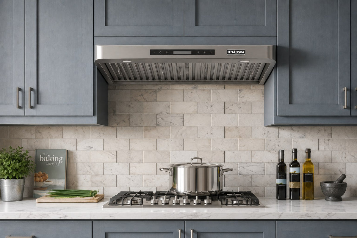 A stainless steel JANSKA range hood installed above a gas cooktop in a modern kitchen with dark gray cabinets and a light stone backsplash.