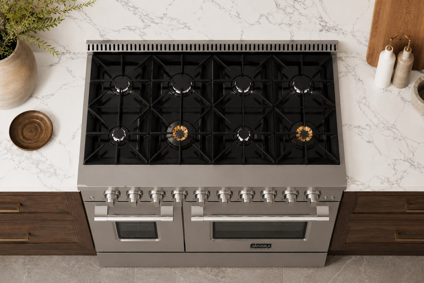 A top-down view of a Janska stainless steel and black gas range with eight burners and two oven doors, set into a modern kitchen with a white marble countertop and dark wood cabinets.